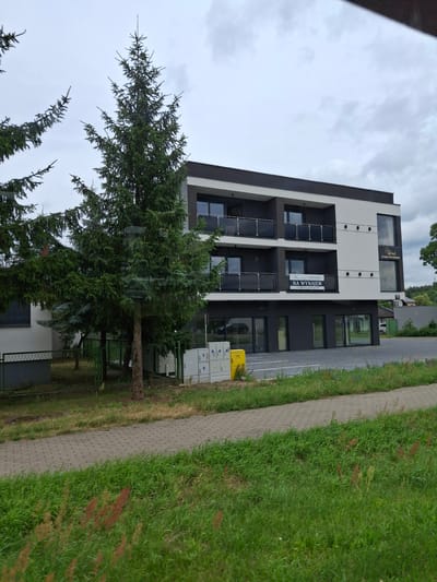 The image shows a modern, multi-story building located in Pila, Poland. The building has a dark grey and white facade with black balconies on the upper floors. A banner advertising "NA WYNAJEM" (For Rent) is visible on the second floor. There are large windows and glass doors at the ground level. In front of the building, there is a paved area with a yellow gas meter box and utility cabinets. Several evergreen trees are situated in front of the building. The scene is captured from a low angle, with a sidewalk and grassy area in the foreground. The sky is overcast.