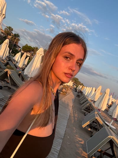A young woman with long blonde hair and fair skin is taking a selfie on a beach. She is looking towards the camera with a neutral expression, her lips slightly parted. Her right shoulder and arm are illuminated by the warm golden light of what appears to be sunset. She is wearing a dark top and a gold necklace. Behind her, a wooden boardwalk stretches along the beach, lined with rows of empty lounge chairs and closed white umbrellas. Palm trees and buildings are visible in the background, suggesting an urban or resort setting. The sky is a clear blue with scattered white clouds. The overall scene conveys a sense of tranquility and the end of a day at the beach, possibly in a location like Manavgat, Turkey, known for its coastal resorts and scenic beauty.