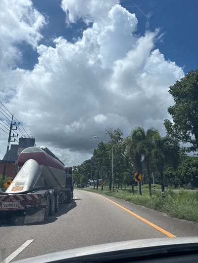 This is a vertical, eye-level shot captured from inside a vehicle, showing a road curving to the right. The sky is predominantly blue with large, dramatic white clouds, suggesting it's daytime, possibly late morning or early afternoon. There are trees lining the road, some with palm fronds, and grassy verges.

The primary object in the foreground is a cement mixer truck, seen from the rear and side. The truck is red and white with multiple axles and wheels. On the back of the truck, there is visible text in Thai script that appears to say "รองพวง". There are also reflective red and white striped markings and a license plate.

The scene appears to be a road in a somewhat rural or suburban area, given the abundance of greenery and the presence of utility poles and wires on the left side of the road. The road itself is paved asphalt with a distinct yellow line marking the lane and a white line on the right edge. A yellow chevron sign indicating a curve is visible on the left side of the road, and another similar sign is further down. The overall impression is of a bright, sunny day with impressive cloud formations.