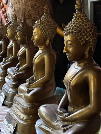 The image displays a close-up, angled view of several golden Buddha statues lying down. The statues are lined up and appear to be made of polished metal, reflecting the light. The texture of their hair is depicted with many small, circular curls. The statues are arranged in a row, with some of them overlapping. In the background, there are hints of a textured surface, possibly a wall or fabric, and some darker, out-of-focus elements. The lighting suggests an indoor setting, possibly a temple or shrine. There are no visible people or clear indications of the time of day or weather. Some reflective plaques with what appears to be text are visible at the bottom of the image, but the text itself is illegible. The overall impression is one of serenity and religious devotion, with the golden statues conveying a sense of opulence and spiritual significance. The location context of Colombo, Sri Lanka, suggests these might be within a Buddhist temple.