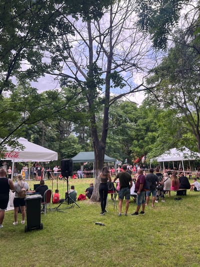 The image depicts an outdoor event in Markovo, Bulgaria. A crowd of people is gathered on a grassy area, likely attending a performance or presentation. There are tents set up, including one with a Coca-Cola logo. Trees surround the event space, with a view of the sky visible through the branches. A speaker system is visible near the front of the audience, suggesting a performance or presentation area. People are seated in chairs facing the direction of the speakers.