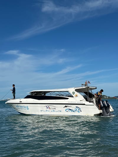 A white speedboat with a dark tinted canopy is on the water under a blue sky with scattered white clouds. Two individuals are visible on the boat. The person on the left, wearing dark clothing, is standing near the bow and appears to be holding a rope. The person on the right, in a blue and yellow shirt and dark shorts, is standing near the stern by the outboard motors and seems to be adjusting some equipment. The boat has text and graphics on its side, including what appears to be Thai script and a logo with the name "MAENAM Beach Marine". The water is choppy with visible waves. In the background, a distant shoreline with buildings and vegetation is visible. The image location is identified as Baan Tai, Thailand.