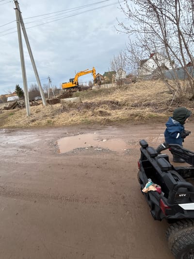 The image depicts a dirt road in the city of Ферма, Russia. A child in a blue jacket and dark hat is standing next to a black UTV. The road is muddy with a large puddle. In the background, there is an excavator, power poles, and vegetation. The sky is overcast.
