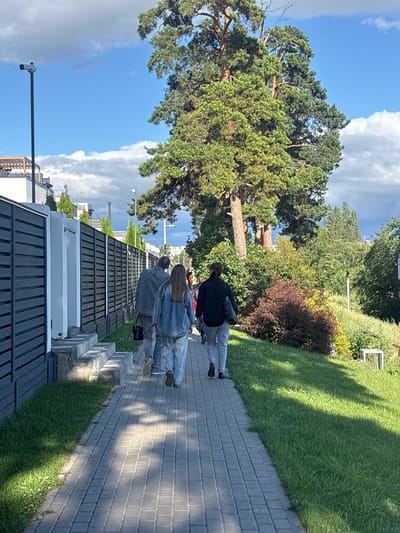 The image shows a paved walkway next to a grassy slope, with several people walking along the path. The walkway is made of gray rectangular pavers, and the grass is a vibrant green, casting long shadows across the scene, suggesting it is daytime. The walkway is bordered by a low, dark fence or wall.

In the background, there is a large evergreen tree with a thick trunk and dense foliage. Beyond the tree, there are other trees and bushes, with a blue sky and white clouds visible above. The overall setting appears to be outdoors in a park-like or residential area.

Three individuals are walking away from the camera, positioned on the walkway. The person in the front is wearing a dark top and light-colored pants. The person behind them is wearing a denim jacket and light pants. The third person, further back, is wearing a dark top and jeans. They are all walking in the same direction.

Notable details include the lighting from the sun, which creates distinct shadows, and the clear blue sky with scattered clouds. The presence of the large tree and well-maintained greenery suggests a pleasant outdoor environment. There is no visible text in the image. The scene appears peaceful and ordinary, with no strong emotional cues conveyed by the subjects. The location context is identified as Volzhskiy Utes, Russia.