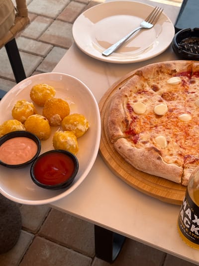 A close-up, overhead shot shows a table set for a meal. The primary subjects are a pizza and a plate of fried appetizers. The pizza, with a thick, golden-brown crust, is topped with melted cheese and what appear to be dollops of mozzarella. It rests on a round wooden board. To the left, a white plate holds a silver fork. In the foreground, a white oval dish contains several golden-brown, fried balls, likely arancini or cheese bites, accompanied by two small black bowls of dipping sauce: one with a bright red sauce, possibly ketchup, and the other with a pinkish sauce. A glass bottle with a yellow label, partially visible in the upper right corner, shows the text "JACK & LYCH". An ashtray with cigarette butts is also visible in the upper left. The setting appears to be an outdoor dining area, suggested by the tiled floor visible in the lower portion of the image. The lighting indicates it could be daytime.