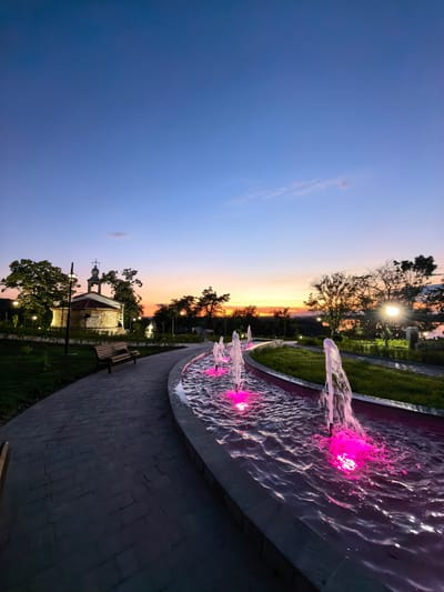 This is a vertical, eye-level, full shot of a fountain with pink-colored lights at dusk in the city of Svishtov, Bulgaria. The fountain is on the right side of the image and is in the foreground. It has multiple streams of water shooting up from it. The water is illuminated by pink lights, giving it a vibrant glow. To the left of the fountain is a paved walkway. Beyond the walkway is a small building with a steeple that resembles a church. There are trees and streetlights around the building. The sky is a deep blue with a hint of orange and pink near the horizon, indicating that the sun has recently set. The overall scene is serene and picturesque.