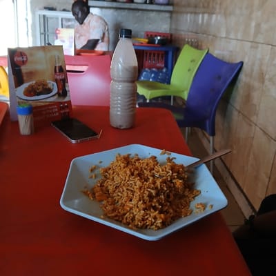 A plate of rice with a spoon on top sits on a red table in front of a half-eaten bottle of chocolate milk. A plastic Coca-Cola ad with a picture of fried rice sits on the red table next to a toothpick holder with a few toothpicks sticking out. Two plastic chairs with blue and green seats can be seen in the background of the image. A man wearing a dark shirt is visible in the very back of the image. The scene appears to be a restaurant. The location is an unnamed restaurant in Kufang, Nigeria, based on coordinates (latitude: 9.863, longitude: 8.867).