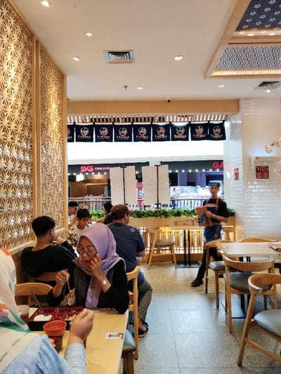 The scene is a restaurant in Telukjambe, Indonesia (latitude: -6.301, longitude: 107.278).  The restaurant features a modern interior with wooden tables and chairs, decorative wooden wall panels, and a ceiling with decorative lighting. Several people are seated at a table, including two women, one man, and a young boy.  The women are eating, and the boy is looking at his phone. A woman is holding a dish in her left hand and her right hand is on her face.  A man is seated next to her, eating.  Two men are seated at a different table.  A man wearing a blue shirt and a blue apron is standing near the table, carrying a food tray in his left hand.  Behind the restaurant is a window that looks out onto a cafe.  The sign above the cafe reads "SKS Game Cafe".  The scene is brightly lit, and the atmosphere is relaxed and casual.