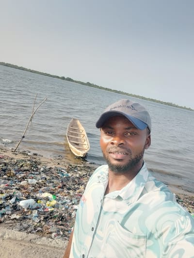 A man is taking a selfie outdoors by a body of water on what appears to be a bright, clear day. He is positioned in the foreground, facing the camera, with a slight smile on his face. He has dark skin, a short beard, and is wearing a light-colored, short-sleeved collared shirt with a wavy, abstract pattern in shades of light blue and white. He is also wearing a grey baseball cap with a blue brim. On the cap, text is partially visible, appearing to be "Ommuso" or similar.

Behind the man, to his left, is a small, light-colored wooden dugout canoe or boat, partially in the water and partially on the muddy/sandy shore. A thin, branched stick or pole is stuck into the ground near the water's edge, slightly to the left of the boat.

The shoreline in the lower left and central-left of the image is heavily littered with a large accumulation of various types of trash, including plastic bags, bottles, and other debris, creating a significant environmental blight. The water beyond the shore is a wide expanse of a lake or river, with gentle ripples visible on its surface. In the far background, across the water, a distant landmass or shoreline with green vegetation (trees or bushes) is visible under a clear, bright sky. The overall impression is of a natural landscape significantly impacted by pollution.