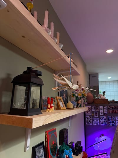 The image depicts a close-up, eye-level, indoor view of a wall with two wooden shelves. The wall is painted a light green.
The upper shelf, made of light-colored wood, has several items. Towards the left, there is a tall, black, square lantern with glass panels, inside which a string of small, white lights is visible. To the right of the lantern, there are two small, red candle holders with yellow flames, and a framed photograph. Further to the right, there is a bouquet of flowers in a vase, and a light brown, round vase.
Suspended from the upper shelf by a thin blue string is a delicate, light-colored origami-like creation, possibly a bird or insect. It is positioned above the middle of the shelf.
The lower shelf, also made of light-colored wood, is positioned below the upper shelf and is supported by white brackets. On this shelf, to the left, is a red book with blue and black text and an image of a face. To the right of the book, there are several framed items, including a black and white photograph. On the far right of this shelf, partially visible, is a dark brown rectangular object.
Below the lower shelf, the wall continues, and on the right side of the image, a section of a refrigerator is visible, covered with many small photographs. The lower portion of the wall below the lower shelf has various objects, including a dark, textured object, a blue and green pixelated figure, and a black camera. There are also two small LED grow lights illuminating the area.
The lighting in the scene is a mix of warm light from the objects on the shelves and the ambient light, with a distinct purple hue emanating from the grow lights on the right side.
The image does not contain any explicit indicators of the city of Chang Phueak, Thailand.