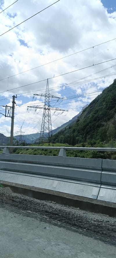 This is a medium shot of a mountainous landscape with power lines in the foreground and midground. The sky is partly cloudy with a mix of white and grey clouds against a blue sky. A large, lattice-style metal power tower dominates the center of the image, with several electrical wires extending from it. To the left, another smaller power pole is visible. In the background, a steep, tree-covered mountain slopes upwards, with lighter patches of rock visible near the summit. A section of a modern highway barrier, made of corrugated metal and concrete, runs horizontally across the bottom third of the image, obscuring the lower part of the landscape. The ground in front of the barrier appears to be a mix of gravel and asphalt. The image captures a scene of infrastructure integrated into a natural, mountainous environment. The location is Golling an der Salzach, Austria.