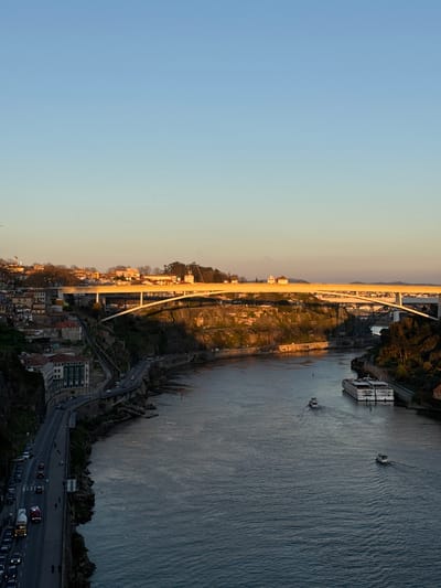 This image captures an elevated, panoramic view of the Douro River in Porto, Portugal, during late afternoon or early evening, characterized by warm, low-angle sunlight.
The sky occupies the upper third of the frame, clear with a gradient from light blue at the zenith to a pale yellowish hue near the horizon, indicative of sunset.
The Douro River flows prominently through the middle ground, its dark blue-grey surface reflecting the sky, with visible ripples and wakes from moving vessels. On the river, one large, white multi-deck passenger cruise ship is docked along the right (north) bank. Two smaller, white motorboats are actively traversing the river upstream, leaving distinct wakes behind them.
The left (south) bank, part of Porto, features a steep, rocky cliff face densely covered with vegetation and numerous multi-story residential buildings with red-tiled roofs, terraced up the hillside. A curved, paved road runs along the base of this cliff, parallel to the river. Numerous passenger cars are visible on this road, some parked and others in motion, heading left (west). A yellow cement mixer truck and an emergency vehicle with blue flashing lights are also visible on the road, traveling in the same direction. Several indistinct figures appear as pedestrians on the sidewalk adjacent to the road.
Three bridges span the river in the mid-ground and background. The most prominent is the Ponte do Infante, a modern white concrete arch bridge, illuminated golden by the setting sun, carrying vehicular traffic. Behind it, further downriver, is the Ponte de São João, a darker metal truss railway bridge, partially obscured by the Ponte do Infante. In the far distance, another large arch bridge, the Ponte da Arrábida, is faintly visible. The right bank primarily shows parts of Vila Nova de Gaia, with scattered buildings and trees illuminated by the setting sun.