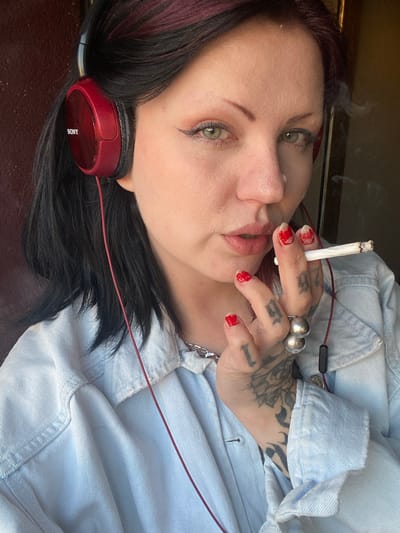 A woman is reclining and holding a lit cigarette near her lips. She has dark hair with red highlights, green eyes, and red nail polish. There are tattoos on her fingers and the back of her hand. She is wearing red Sony headphones and a light blue denim shirt. A silver necklace is visible. The location is not identifiable from the image.