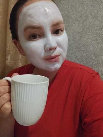 A person with a white facial mask is holding a white, ridged mug while wearing a red shirt. The background includes a light-colored wall and a partially visible gold curtain. The image appears to have been taken indoors, possibly in Kuyeda, Russia.