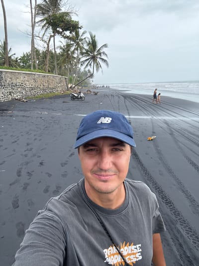 A male subject is depicted in a selfie on a black sand beach in Beraban, Indonesia. He occupies the foreground, wearing a blue baseball cap with a white "NB" logo and a dark grey t-shirt with "PARADISE MADNESS" text. The subject has light skin and a slight smile.
The background reveals a volcanic black sand beach marked by numerous tire tracks and footprints. To the left, a dark stone retaining wall parallels the coastline, behind which stand multiple palm trees and tropical vegetation. A white scooter is parked on the sand near the wall. Further down the beach, several dark objects resembling small fishing boats or sleds are visible.
In the mid-ground to the right, two individuals, an adult and a child, walk along the wet sand near the ocean. The sea extends to the horizon, where waves are breaking. The sky above is overcast. A small, colorful object, appearing to be an offering or discarded item, lies on the sand to the right of the primary subject.