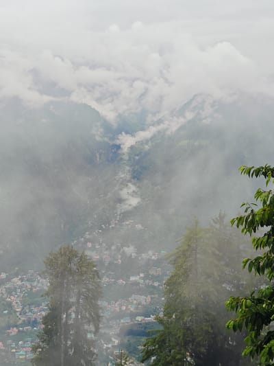 The image shows a mountainous landscape with a town nestled in the valley. The sky is overcast with clouds, and there is a light mist or fog obscuring some of the details. In the foreground, there are several large evergreen trees. Beyond the trees, the town of Prini, India, is visible, with a dense collection of buildings spread across the hillsides. The buildings are of various colors, including white, pink, and blue. A winding road can be seen leading through the town. Further in the background, the mountains rise steeply, covered in dense vegetation and more clouds. The overall scene conveys a sense of natural beauty and a remote, mountainous settlement.