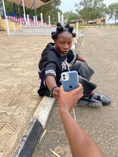 A young Black woman with braided hair sits on the edge of a paved area next to a curb in Zawan, Nigeria. She is looking down at a blue smartphone held by an unseen person. The woman is wearing a black hoodie with white stripes on the sleeves and dark pants. She has a black beaded bracelet on her left wrist. The background shows a stadium with red seats, trees, and buildings, suggesting an outdoor public space such as a school or sports complex. The ground is a textured paving stone near the curb, transitioning to asphalt further away. The lighting indicates it is daytime, possibly overcast.