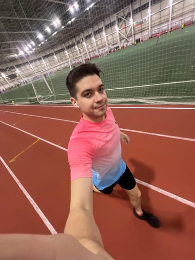 The image shows a young man posing for a selfie on an indoor running track. The photo is rotated 90 degrees clockwise, so viewed upright, the man is standing on a red track with white lane markings. He is positioned slightly off-center, facing the camera with a neutral expression and a faint smile.
The man has short, dark hair, neatly styled, and prominent dark eyebrows. He is wearing a short-sleeved athletic shirt that transitions from bright pink on top to light blue at the bottom. He also wears black athletic shorts and a black athletic shoe on his right foot, which is slightly raised as if in mid-step or preparing to move. A white wireless earbud is visible in his right ear. His right arm is extended into the foreground from the right edge of the frame, presumably holding the camera, while his left arm is bent at the elbow.
The background reveals a spacious indoor sports facility. Immediately behind the man and the running track is a tall green netting, likely separating the track from a larger sports field, possibly for soccer or another indoor sport. Beyond the netting, the facility features a high ceiling with numerous artificial lights and intricate metal support beams, indicative of a large modern arena or gym. Small, indistinct figures of other people can be seen in the far background on the green field, suggesting an ongoing activity or training session. The overall lighting is bright and even, consistent with an indoor environment.