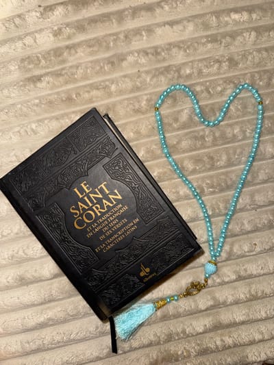 An overhead view in Wattrelos, France, displays a black hardcover book and a string of light blue prayer beads on a ribbed, light-beige fabric surface. The book, titled "LE SAINT CORAN" in gold lettering with French translation and Latin transcription, is positioned diagonally with its spine facing the upper-left. Embossed decorative patterns adorn its cover. To the right of the book, the prayer beads, consisting of pearl-like beads and occasional gold accents, are arranged in a heart shape. A larger, embellished bead leads to a light blue tassel with gold components, which extends towards the bottom-right, partially overlapping the book's lower edge. No individuals are present.