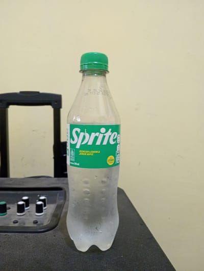 A clear plastic bottle of Sprite, a carbonated soft drink, is positioned in the center of the frame, against a light beige wall. The bottle has a green cap and condensation on its surface. The label on the bottle is predominantly green with white text that reads "Sprite" in large letters, followed by smaller text in Indonesian indicating it is flavored with lemon and lime. The bottle is standing on a dark, textured surface, which appears to be some sort of equipment with several black and white knobs and a green knob visible in the foreground to the left. To the left of the bottle, a portion of a black, structured object, possibly part of a piece of equipment or furniture, is visible. The lighting is even, suggesting an indoor environment, and there is no indication of the city of Keudekrueng, Indonesia in the image itself.