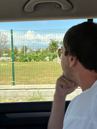 The image depicts a person viewed from behind, looking out of a car window. The person has dark hair, is wearing sunglasses, and has their chin resting on their fist, with a ring on their finger. Outside the window, a green metal fence is visible, with a grassy field behind it. Trees and some buildings can be seen in the distance, under a blue sky with white clouds. The scene appears to be in a semi-rural area, consistent with the outskirts of Kruševica, Montenegro.