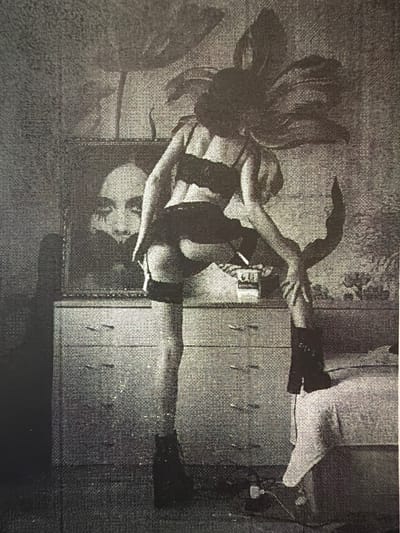 The image depicts an indoor scene, likely a bedroom or dressing area. A person is standing in the center, facing away from the camera and to the right. They are wearing what appears to be lingerie, including a bra, stockings, and boots. The person is holding onto their leg, possibly adjusting their attire.

In the background, a dresser is visible with several drawers. On top of the dresser, there is a framed picture or painting featuring a close-up of a face with striking eyes and dark makeup. A pack of cigarettes and a single cigarette are also present on the dresser.

The environment appears somewhat cluttered and dimly lit. The walls have a textured pattern. A bed is partially visible on the right side of the frame. The overall impression is one of a private, perhaps intimate, setting.

There is no indication in the image that places the scene in the city of Svilengrad, Bulgaria.