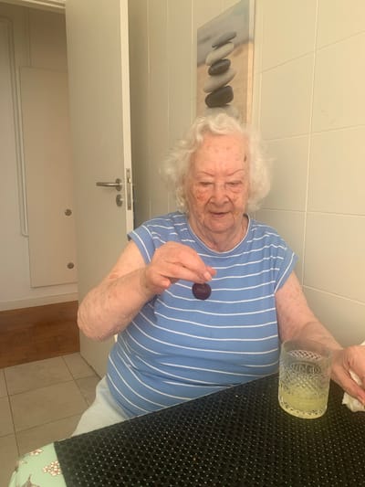 An elderly woman with white hair and fair skin, wearing a blue and white striped t-shirt, sits at a table holding a dark purple plum. A glass of yellow liquid, likely juice, is on the table in front of her, to her right. A white napkin is also visible on the table, partially obscured by her hand. The background consists of a tiled wall and a partially open door leading to another room. A framed picture of stacked stones hangs on the wall. The scene appears to be indoors, possibly in a kitchen or dining area. There are no visible indications of the location being in Lisbon, Portugal.
