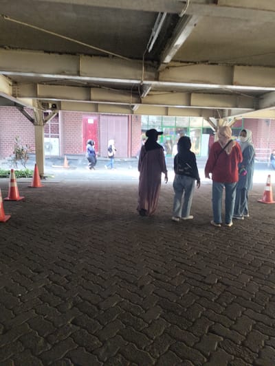 The image depicts an outdoor scene, possibly an underpass or a covered walkway, with brick walls and concrete beams visible. The ground is paved with dark interlocking bricks laid in a herringbone pattern. Several individuals are present, mostly women wearing headscarves, walking or standing in groups. There are orange traffic cones placed on the ground, suggesting an area that might be cordoned off or managed. In the background, there appears to be a building with large doors and windows. The lighting suggests it is daytime, but the covered nature of the area casts shadows. The scene is likely in a public urban space within Bekasi, Indonesia, given the clothing styles and architectural elements.