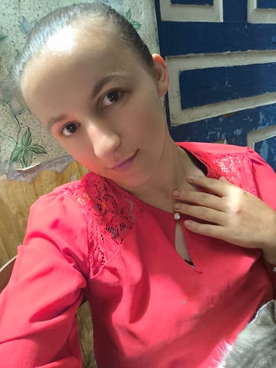 A woman is taking a selfie in Anastasivka, Ukraine. She is wearing a coral pink blouse with lace detailing on the shoulders and a keyhole neckline. Her hair is pulled back, and she has a hand resting on her chest. She is looking at the camera with a slight smile. The background appears to be a room with a blue and white patterned wall and a floral wallpaper. The lighting suggests it is daytime, and the overall mood is casual and relaxed. A hint of a grey cat can be seen on the right side of the frame.