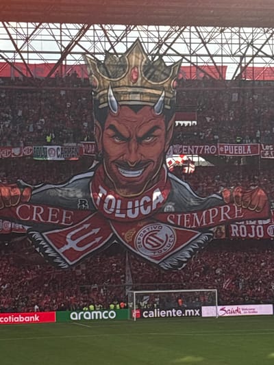 The image captures a vibrant scene within a stadium, possibly in Toluca, Mexico, given the presence of the "TOLUCA" lettering on a large devil-themed banner. A devil character with a crown and horns is centrally featured on the banner, wielding a trident and wearing a scarf, with the phrases "CREE TOLUCA" and "SIEMPRE ROJO". The backdrop shows a densely packed crowd, presumably fans attending a sporting event. Stadium architecture is visible behind the banner. The edge of the soccer field is lined with advertisements, including "caliente.mx," "aramco," and "cotiabank." Another advertisement is for "Saudi - Welcome to Arabia". There are additional signs, including "CENTRO77RO PUEBLA LOS" and "OLLI CARMEN VALNA".