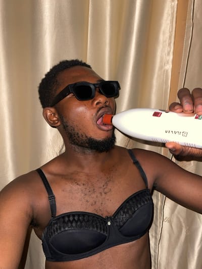 A dark-skinned person, wearing sunglasses and a black bra, lies on a tan satin-like surface. A hand holds a white device labeled "BRAVIA" with an orange tip near their open mouth and extended tongue. The person has a beard and short, dark hair. The scene is captured from a slightly angled perspective, with the background surface appearing smooth and reflective.