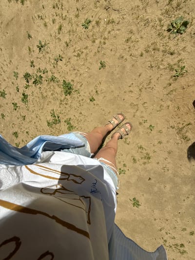 The image shows a person's legs and feet from a high-angle perspective, standing on sandy ground with sparse green weeds. The person is wearing light-colored sandals with two straps and distressed denim shorts. A blue and white striped garment, possibly a shirt or dress, is also visible, along with a white top featuring a sketch-like drawing in gold. The sandy ground shows imprints of footprints and some dry leaves. The lighting suggests it is daytime, with shadows cast on the ground. The overall setting appears to be an outdoor, possibly natural, environment. No specific event or activity is clearly depicted beyond standing. There is faint blue cursive text on the white top, possibly a name, and bold gold lines forming part of a drawing.