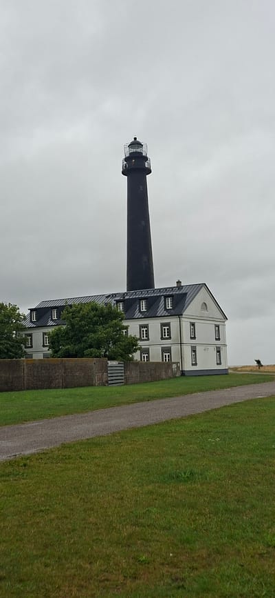 The image depicts the Sääre lighthouse and associated buildings in Sääre, Estonia, under a cloudy sky. The lighthouse is a tall, dark cylindrical structure with a lantern room at the top. In front of the lighthouse is a long, white building with a dark, sloped roof featuring dormer windows. A stone wall runs horizontally across the lower portion of the image, separating the manicured lawn and gravel path from the buildings. Trees are visible behind the stone wall and to the left of the white building. In the background, a person is walking or standing on a grassy area near the sea, with sparse vegetation and a distant, wind-swept tree. The overall environment is coastal and appears somewhat bleak due to the overcast weather.