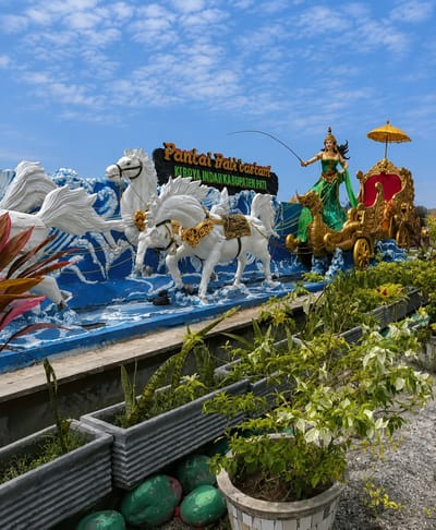 An outdoor, eye-level, medium shot captures a large, colorful public art installation featuring a statue of a woman in a green dress in a golden chariot pulled by four white horses. The installation is set against a bright blue sky with scattered white clouds. A yellow umbrella is positioned over the chariot. A sign behind the installation reads "Pantai Pari castam KEROYA INDAH KABUPATEN PATI". The art piece is situated in front of a blue wave-like structure. In the foreground, planters filled with green plants are arranged in rows, and large green painted spheres are placed on the ground. The location appears to be a coastal area in the city of Pati, Indonesia.