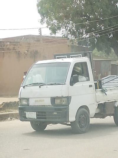 A white Daihatsu Hijet utility pickup truck is positioned on an asphalt road in Hotoro South, Nigeria. The vehicle is oriented facing left, with its front bumper and cab visible. The truck features a black plastic front bumper, a missing headlight in the right housing, and a present headlight in the left housing. The front grill displays a partial "HIJET" logo. A Nigerian license plate, predominantly white with black characters, is partially visible and identifies the vehicle as from Kano state.
Inside the cab, a driver is visible, wearing a light-colored cap or hat. At least one additional passenger, also in light-colored attire, occupies the front passenger seat. The truck's cargo bed contains multiple metallic frames or sheets, secured with green straps or rope. Above the cab, a black roof rack assembly holds two rectangular silver-colored objects.
The immediate environment consists of the asphalt road. In the background to the left, a light brown/beige building wall with visible discoloration and wear is present. Above the wall, multiple utility wires are strung across poles. A large tree with green foliage dominates the upper right background. Further right, a corrugated metal structure is partially visible behind the truck. The sky is overcast or hazy, indicating diffused daylight. A person in an orange top is partially visible behind the truck on the left side, near the building wall.