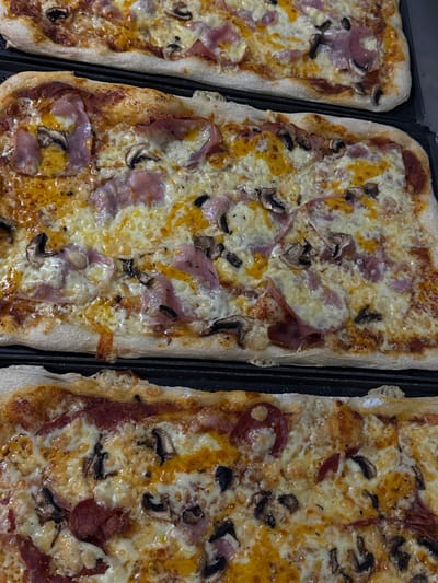 The image shows three rectangular pizzas placed on a dark baking tray. The pizzas are topped with melted cheese, ham, and mushrooms. The crusts are golden brown and appear to be crispy. The pizzas are arranged side-by-side, filling most of the frame. The lighting is somewhat dim, suggesting an indoor setting. The overall impression is of freshly baked food, ready to be served. There is no visible text in the image.