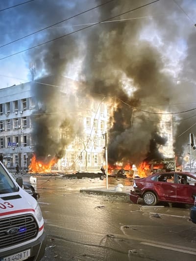 The image depicts a dramatic scene of a building on fire, with thick black smoke billowing into the sky. Flames are visible at the base of the building, engulfing the lower floors and street level. Several cars are present in the foreground and on the street, one of which is a red sedan with its rear end damaged. Another vehicle, appearing to be a white emergency van with red markings and the number "03" visible, is parked in the lower left corner of the frame. The environment appears to be an urban street with power lines overhead. The building itself is multi-story with numerous windows. The ground is wet, possibly from firefighting efforts or debris. A person in dark clothing is visible near the burning building, and another figure is seen on the roof of the building amidst the smoke. The overall scene suggests a catastrophic event with significant damage and active firefighting or rescue operations. The provided location of Pori, Finland, is not directly discernible from the visual elements of the image itself.