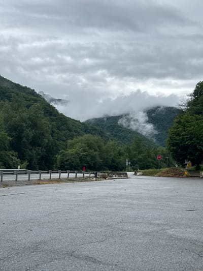 The image depicts a roadside scene in the vicinity of Nizhnyaya Teberda, Russia, characterized by a cloudy, overcast sky and lush green mountains. In the foreground, a paved road or parking area with visible cracks and patches occupies the majority of the frame. To the left, a guardrail with metal posts and horizontal bars separates the paved area from a sloping embankment covered in vegetation.
Further back, the road appears to curve and merge with another paved surface. A stop sign is visible on a pole in the middle distance. The background is dominated by heavily forested mountains, with some areas shrouded in mist or low-lying clouds, suggesting a damp or foggy atmosphere. The trees are dense and varied in shade of green. A few scattered structures or elements of infrastructure, such as concrete barriers and what might be a small building or kiosk with a yellow sign, are present on the right side of the road. No individuals are clearly visible in the scene. The overall impression is one of a rural, mountainous road under a somber sky.