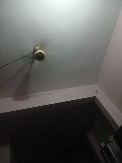The image shows a ceiling with a light fixture and a hook on the wall. The ceiling is painted white and appears to be the interior of a room. The light fixture is white with a brass-colored base and a dark bulb. A black wire is connected to the light fixture and extends to the left. The wall below the ceiling is painted a light pink color. On the pink wall, there is a small, metal hook. The rest of the image is dark, suggesting the room is dimly lit, except for the light from the fixture. This scene is located in Peshawar City Tehsil, Pakistan.