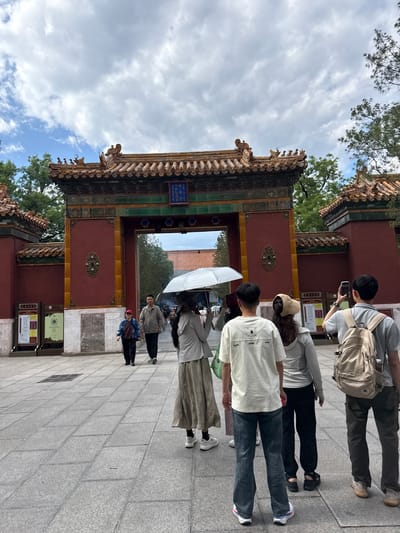 A group of tourists stand in a courtyard in front of an ornate, traditional Chinese gate in Dongcheng District, China. The gate is painted red with yellow accents and features intricate roof decorations and a blue plaque with Chinese characters. The ground is paved with gray tiles. Several people are visible, including a couple walking towards the gate, one holding an umbrella, and a group of younger individuals facing away from the camera, some holding phones. The sky is partly cloudy.