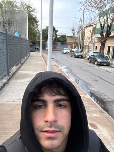 The image is a selfie taken from a low angle on a street in Avellaneda, Argentina. The subject, a young man with dark hair, is wearing a black hooded jacket and the hood is up. He is looking directly at the camera. In the background, there is a sidewalk with a grey fence on the left and a street with several parked cars. The street appears wet, possibly from recent rain. Buildings line the right side of the street. Overhead power lines are visible. The sky is overcast.