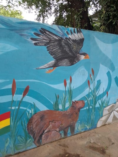 The image shows a mural in Villa Hayes, Paraguay, depicting a bird of prey flying above a family of capybaras in a marshy environment. The bird has dark brown and gray feathers with orange legs and beak. The capybaras are brown, with an adult and a smaller juvenile. The backdrop is a blue sky with white wave patterns. Cattails grow around the capybaras. The Bolivian flag is painted in the corner. The mural is on a concrete wall under trees.