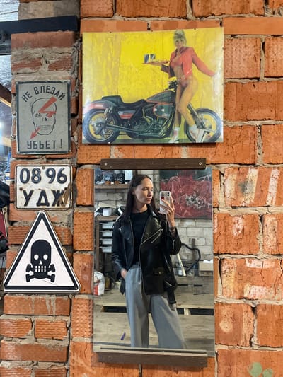 The image shows a brick wall decorated with various items. These include a poster of a woman on a motorcycle, a mirror reflecting a woman taking a selfie, two Russian-language warning signs, and a triangular sign with a skull and crossbones. The location is Oktyabr'skii, Russia, inferred from the Russian writing on the license plate.