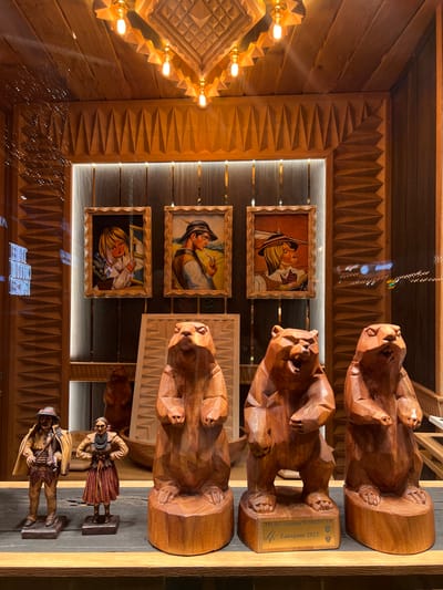 The image shows a display in a shop window, likely in Zakopane, Poland, given the inscription on the trophy. The display features wooden carvings of animals and people, along with framed artwork. In the foreground, there are three large wooden carvings of bears, with the middle and right bears positioned to the right of the image. The bear on the right has a plaque at its base that reads "FIS Ski Jumping World Cup Zakopane 2023". To the left of the bears are two smaller wooden figurines of a man and a woman, dressed in traditional attire. Behind these figures, three framed paintings are displayed, each depicting a person in traditional clothing. The artwork and carvings are set against a backdrop of wooden paneling, with decorative carvings adorning the walls. The ceiling features wooden paneling and decorative lighting. The overall impression is of a display of traditional Polish folk art and memorabilia.