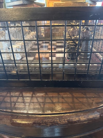 The image shows a close-up view of a display case containing a chess set behind a black metal grate. The chess pieces appear to be intricately designed, with some pieces being light-colored and others dark-colored. A price tag with the number "2900" is visible on a small wooden stand in front of the chess set. The background shows stacks of books. The display case is situated on a dark wooden surface, which reflects the shadow of the grate. The setting is indoors, likely a shop or museum, with warm lighting. There are no people visible in the image. The location is not specified in Tongzhou District, China.