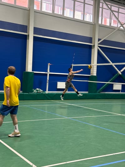 The image depicts an indoor volleyball game in Sinyavino, Russia. Two individuals are visible on a green court marked with white and blue lines. One player, wearing a gray tank top and shorts, is in mid-air, jumping to hit a volleyball. The ball is positioned above and in front of him, with his arm extended towards it. He is wearing white socks and athletic shoes. Another person, wearing a bright yellow t-shirt and blue shorts, stands on the court facing away from the viewer. The setting is an indoor sports facility with a high ceiling and large windows on one side, revealing an exterior view of buildings. The walls of the court are a vibrant blue, and a net is visible across the middle of the court. The lighting suggests it is daytime. The overall atmosphere appears to be one of active sports participation. There is no visible text in the image.