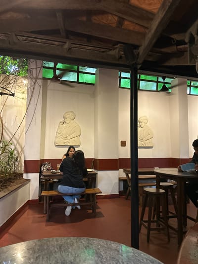 This image depicts a rustic and traditionally-styled cafe or restaurant interior, likely in Kochi, India, given the location context. The lighting suggests it is either evening or a low-light indoor environment. The scene is captured from a slightly elevated perspective, looking down into a seating area.
In the foreground, a person with long dark hair, wearing a black top, light blue jeans, and white sneakers, is seated at a dark wooden table. They turn their head towards the viewer, offering a subtle smile. A patterned bag rests on the chair beside them. Another individual is partially visible in the upper right, seated at a different wooden table, with only their lower body and sandals showing.
The decor includes white walls prominently featuring two large, light-colored bas-relief sculptures. These carvings depict a seated figure, possibly a sage or elder in traditional attire, adding a cultural element to the space. Multiple windows with green-tinted glass panes and dark frames are visible along the far wall, providing some filtered light and a view of outdoor foliage, including bare branches.
The structural elements of the space are notable, with heavy dark wooden beams forming the ceiling structure and a prominent black horizontal beam crossing the mid-ground. The floor is a reddish-brown color, likely tiled or concrete. The overall activity involves people seated at tables, suggesting dining or socializing. Small, indecipherable plaques or labels are visible on a wall and a table.