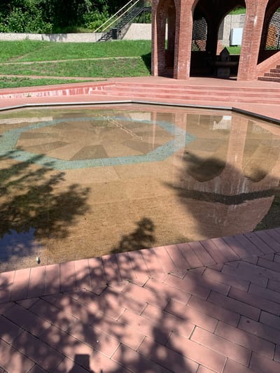 A shallow, decorative fountain with a geometric pattern is centrally located in a sunlit outdoor area. The fountain's base is tiled with large, light brown granite slabs arranged in a star-like pattern, intersected by green tiled bands. Tree shadows are cast across the water and the surrounding reddish-brown brick-paved walkway. To the left, a grassy slope rises, with a stone wall and a black metal staircase leading upwards. To the right and background, an arched brick structure, possibly a gazebo or pavilion, is visible, with red brick columns and tiered seating. The scene appears to be in a public park or recreational area. The specific location in Arbuzov-Baran, Russia, is not discernible from the image content alone.
