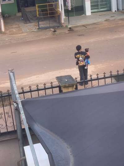 In the city of Shagamu, Nigeria, a person stands holding a child in their arms. The individual is wearing a black t-shirt with a graphic design and black pants. The child, facing away from the camera, is wearing an orange top and blue pants. They are standing on a dirt road, with a black metal fence in the foreground and buildings in the background. The overall scene suggests a residential or commercial street environment.
