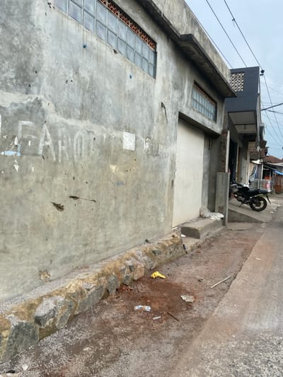 The image shows the side of a concrete building with a large, white garage door. The building has a row of windows made of small glass squares and a concrete foundation. The building has some damage to the exterior, including cracks and missing sections of concrete. There is a row of large rocks along the base of the building and a small, yellow plastic bag lying on the ground in front of it. The building is located on a narrow, paved street. There is a motorcycle parked on the street to the right of the building. The location is a street in the city of Margaasih, Indonesia, based on coordinates (latitude: -6.957, longitude: 107.549).